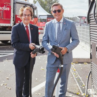 Saubermacher zeigt Ladeinfrastruktur für E-POWER-Lkw – © Saubermacher/eap.at Zwei Vertreter stehen mit Ladesteckern neben einer Saubermacher-Ladestation, im Hintergrund ist ein E-POWER-Lkw mit Hinweis auf elektrischen Betrieb zu sehen. © Saubermacher/eap.at