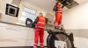 Zwei Servicemitarbeiter von Saubermacher Outsourcing in roter Arbeitskleidung führen eine Lüftungsrohrinspektion in einem Küchenbereich durch. Einer steht auf einer Trittleiter und inspiziert mit einer Kamera ein offenes Lüftungssystem in der Decke, während der andere technische Geräte am Boden bedient. Diese Inspektionen sind seit Oktober 2024 gemäß § 13 der Arbeitsstättenverordnung gesetzlich vorgeschrieben​. Fotorechte: Saubermacher