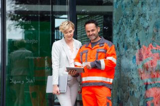 Arbeiter und Angestellte©Majland Eine Angestellte mit Laptop und ein Arbeiter in orangefarbener Schutzkleidung betrachten gemeinsam ein Tablet vor einem Unternehmensgebäude. Die Szene steht für Zusammenarbeit zwischen Büro und operativem Bereich. ©Majland