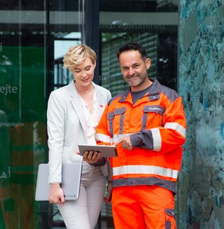 An office worker with a laptop and a worker in orange protective gear are looking at a tablet together in front of a company building. The scene symbolizes collaboration between the office and the operational side of the business. ©Saubermacher_Majland
