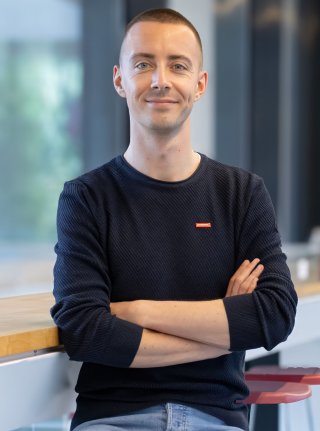 Junger Mann mit kurzen Haaren und verschränkten Armen sitzt in einem modernen Büro vor einem Tisch und lächelt in die Kamera – Fotocredit: Martina Majland