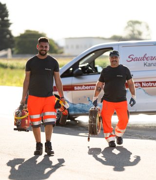 Zwei Mitarbeiter von Saubermacher in orangefarbener Warnkleidung und schwarzem Shirt gehen mit professioneller Ausrüstung zur Kanalreinigung auf einen Einsatz zu. Im Hintergrund ist ein Saubermacher-Fahrzeug mit der Aufschrift „24h Notruf“ zu sehen. Die Szene vermittelt Kompetenz und Einsatzbereitschaft bei Problemen wie Kanalverstopfungen oder Wurzeleinwuchs. Fotocredit: Saubermacher