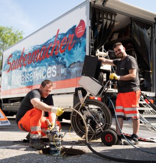 Zwei Mitarbeiter von Saubermacher führen mithilfe modernster Technik eine Kanalinspektion durch. Einer arbeitet direkt an einem offenen Schacht, während der andere ein Kamerasystem bedient. Im Hintergrund ist ein Servicefahrzeug mit der Aufschrift „Saubermacher Kanalservices“ zu sehen. Die Szene steht exemplarisch für professionelle Kanaldienstleistungen wie Dichtheitsprüfung, Rückstauklappenwartung und Kanalsanierung. Fotocredit: Saubermacher