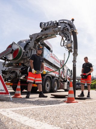 Zwei Saubermacher-Fachkräfte arbeiten mit einem Spezialfahrzeug zur Kanalwartung an einem offenen Kanalschacht auf einem Parkplatz. Der moderne Spül- und Saugwagen ist im Einsatz, um die Infrastruktur zuverlässig instand zu halten und Verunreinigungen zu vermeiden. Sicherheitspylonen sichern den Einsatzbereich ab.